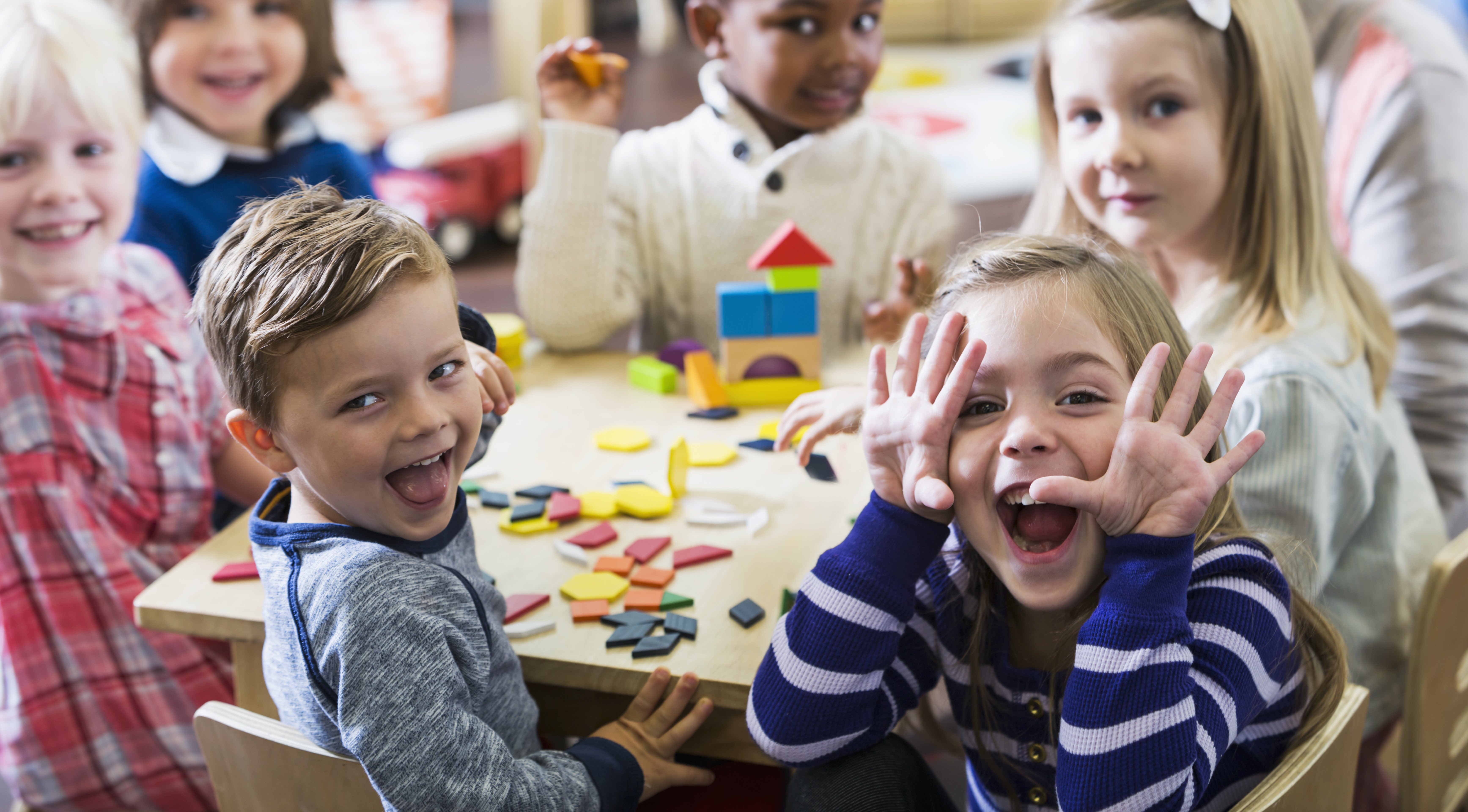 A multiracial group of preschoolers or kindergarteners having fun in the classroom. Six children are sitting around a little wooden table playing with colorful wooden block and geometric shapes. The playful little girl in the foreground is making a silly face at the camera.