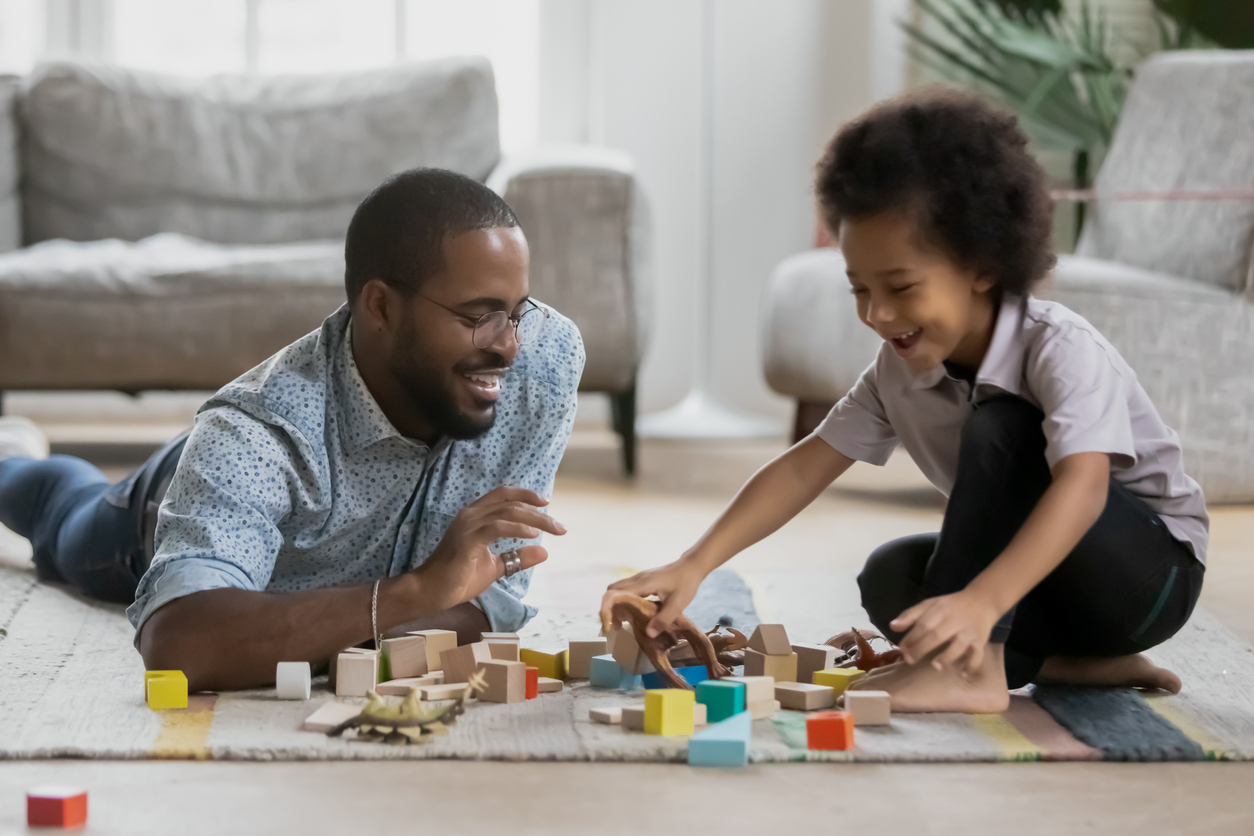 Excited young african American father and small son sit on floor in living room construct with wooden blocks together, playful biracial dad and little boy child have fun play with building bricks toys